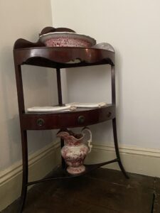 This Sheraton-style corner washstand sits beside a bed in the bedchamber at the Mount Vernon Hotel. Carved into it is a large hole to fit a washbasin and served as a convenient place for guests to wash up and ready themselves for the morning. 